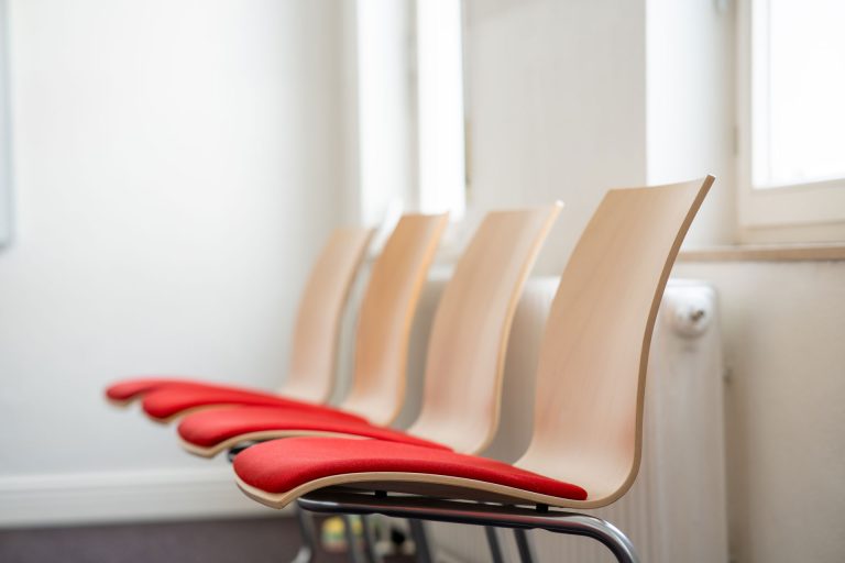 Moderner Wartebereich der Praxis für Kinder- und Jugendpsychiatrie von Andrea Fleßner-Polychronidis in Hannover-Kirchrode. Mehrere helle Holzstühle mit roten Sitzpolstern stehen nebeneinander in einem lichtdurchfluteten Raum.